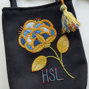 Produktbild: En liten svart broderad väska med en tofs som hänger och en broderad blomma.